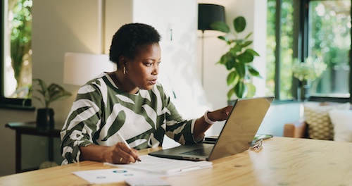 Woman at dining room table working on laptop with papers nearby.