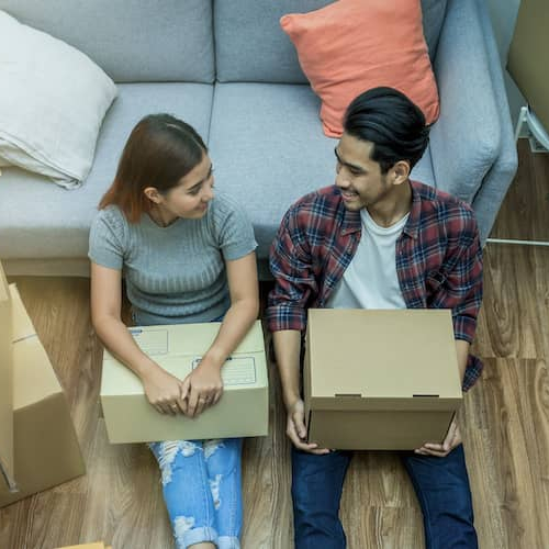 A young Asian couple smiles as they unpack cardboard boxes in a warmly lit living room, creating a cozy scene of settling into their new home.