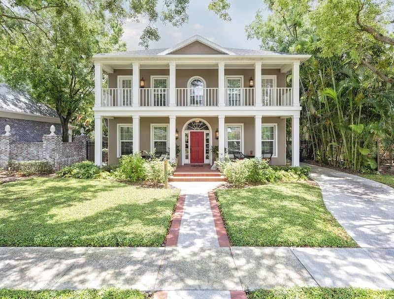 Two story tan French Colonial home with white columns featuring covered porches on both levels and a red door.