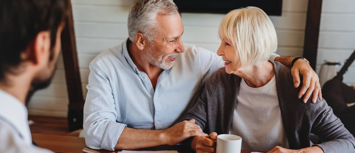 Older couple smiles at each other while drinking coffee and chatting with a real estate agent or advisor.