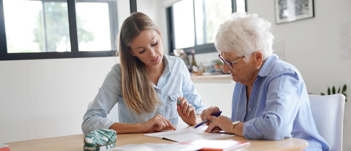 Woman helping her elderly mother fill out paperwork in a sunny kitchen.