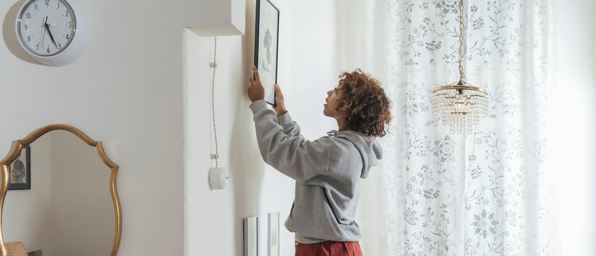 Young woman with curly hair hands up a painting in her apartment.