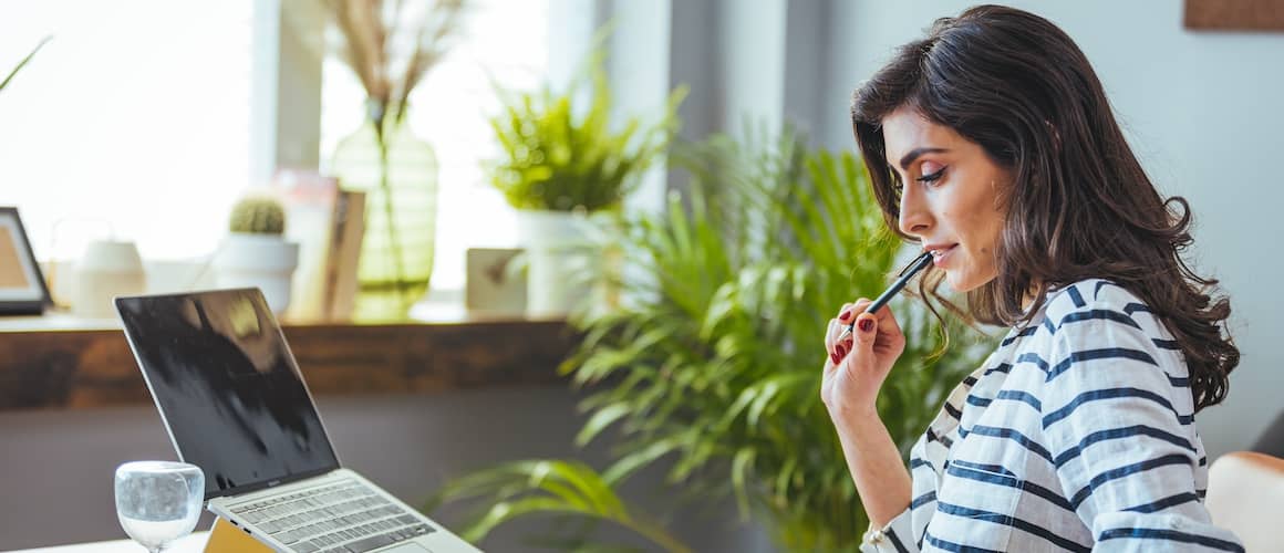 Woman Sitting at desk in striped shirt looking at a laptop and holding a pen.