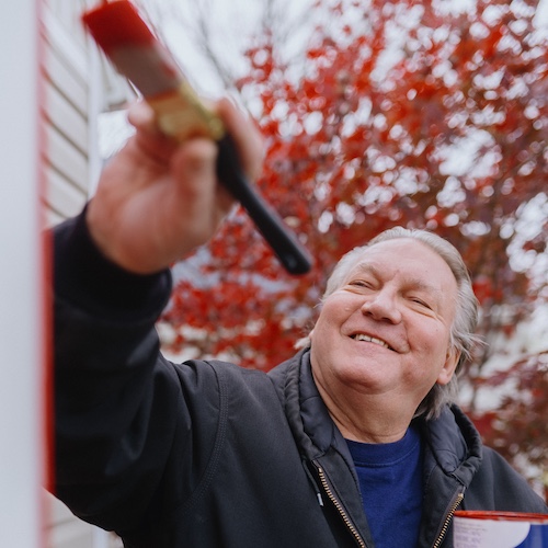 Man smiling and painting exterior of home in red with a paintbrush.