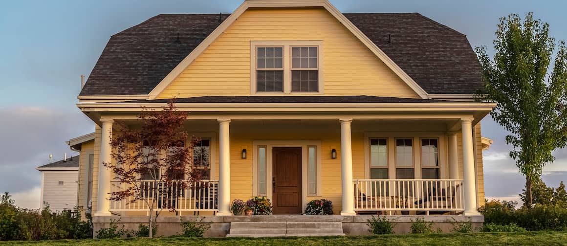 Traditional pale yellow house with Greek columns and wrap-around porch at dusk.