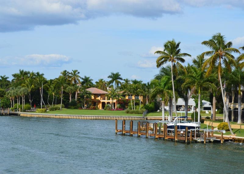 View of luxury waterfront home with dock with boat  dotted with palm trees along property.