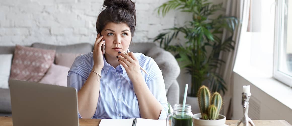 Woman on cell phone at home office desk with open laptop in front of her.