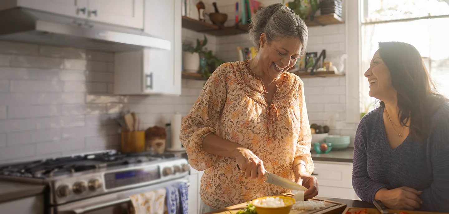 Dos mujeres hispanas, una madre mayor y una hija, cocinando en la cocina y riéndose.