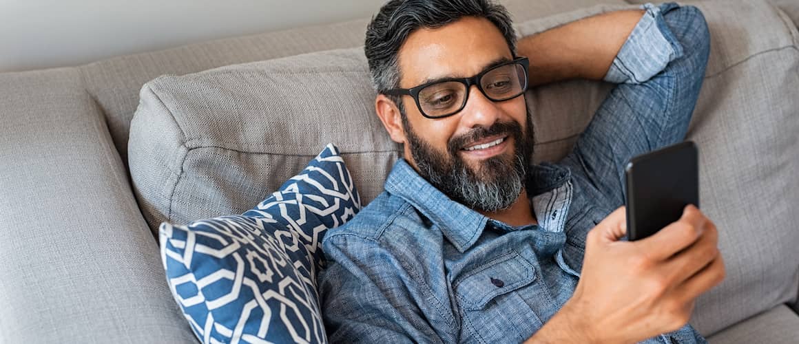 Image of man holding cell phone on couch, smiling at screen.