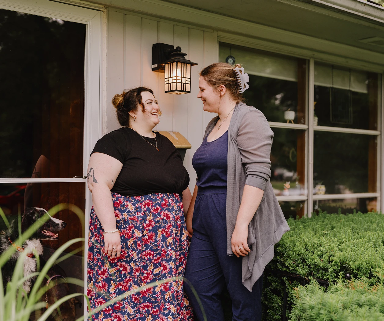 Two women look lovingly at each other in front of their house.