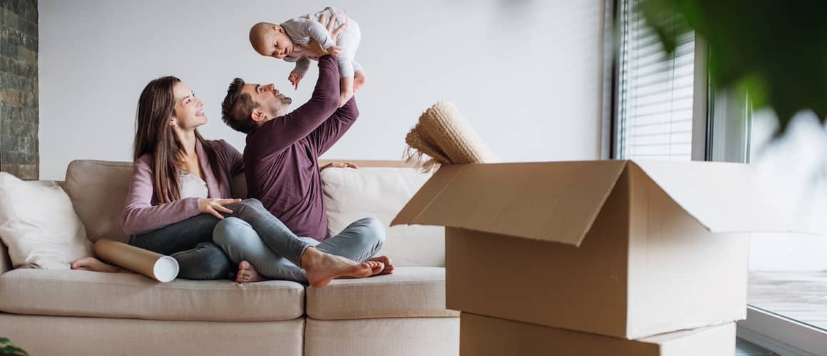 Young couple holding curious baby sitting on the couch while unpacking cardboard boxes.