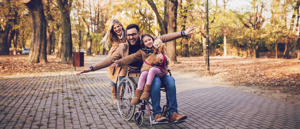A father in a wheelchair with his wife and daughter in a park setting.