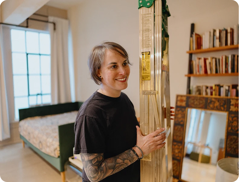 A smiling woman holding a ladder in a room with a bookshelf and a large window.