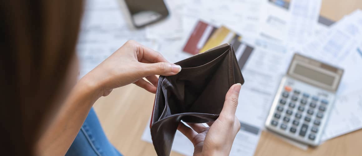 Woman sitting on floor opening empty wallet, with bills spread out in front of her and calculator, phone nearby.