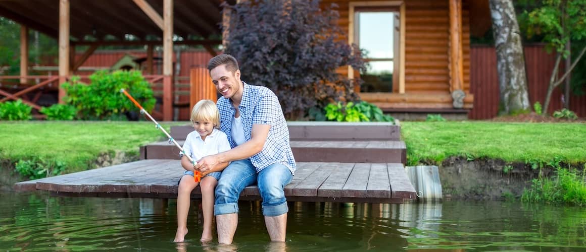 Dad and son sitting on the dock fishing with feet in the water in front of a wooden cabin.