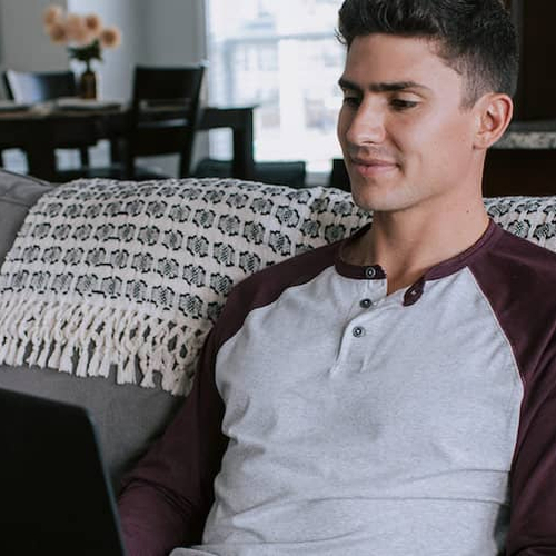 A young man sitting on his home couch looking at something on his laptop.