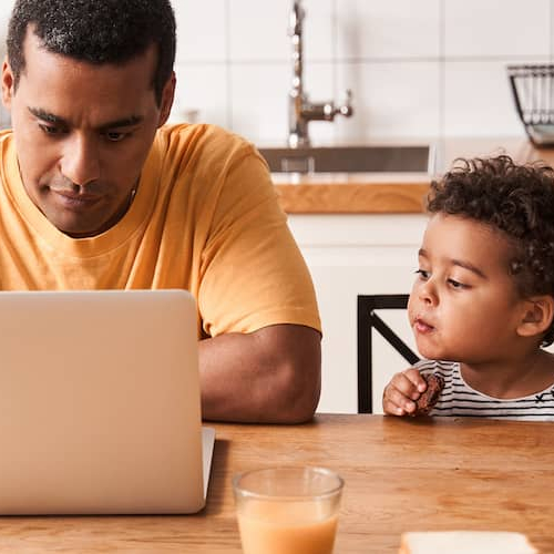 A man working in his kitchen while his child sitting beside is looking at it.