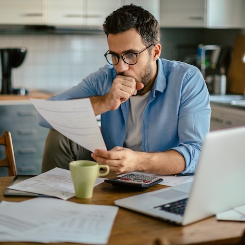 Man looking over paperwork in his kitchen with a calculator and laptop beside him.