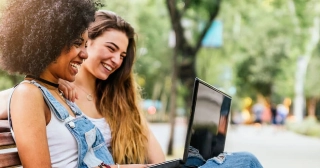 A young Black woman in denim overalls and her friend, a young white woman with long hair, smile while looking at a laptop together on a park bench. 