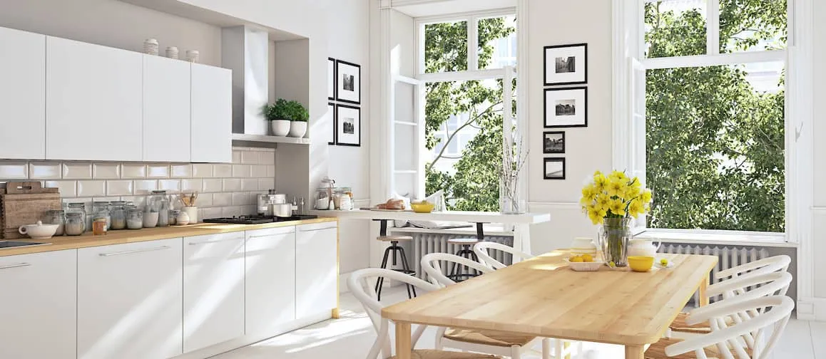 A clean, white kitchen in Scandinavian interior design style. Two open windows show a green tree outside. There are yellow daffodils in a vase on the white wood kitchen table.