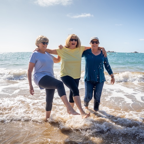 Three senior women laughing and dancing in shallow waves on the beach.