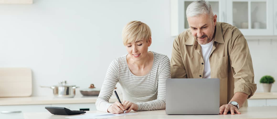 A middle-aged couple at a kitchen island using a computer and calculator, potentially managing finances or planning for a home purchase.