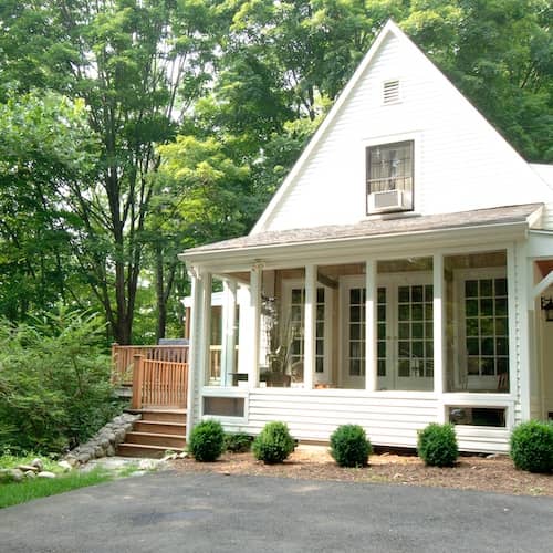 White home with large covered porch surrounded by forested lot.