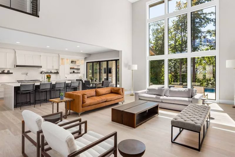 Open concept living room with a brown leather couch floating between bar style open kitchen. The home has high ceilings with floor to ceiling windows looking out onto wooded lot.