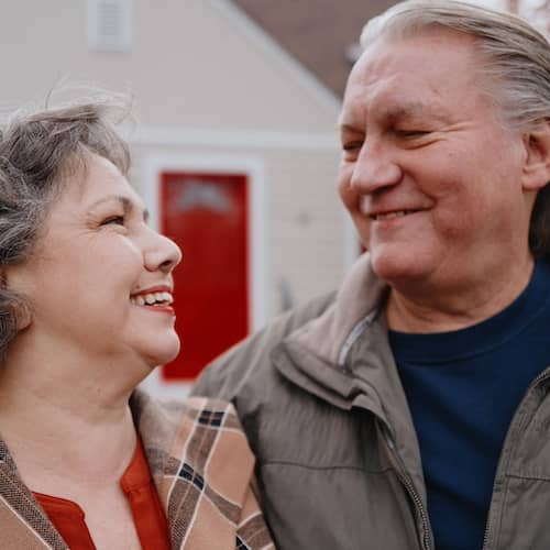 Older couple smiling at one another in front of home.