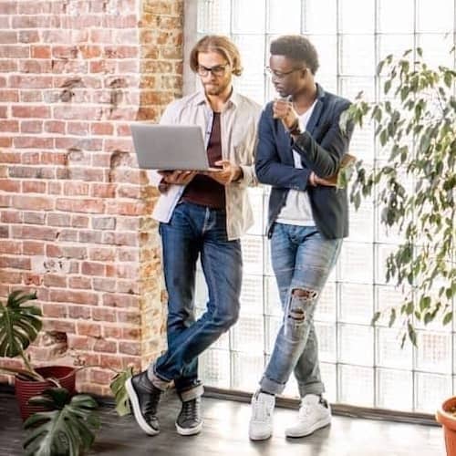 Couple looking at open laptop standing up in loft with houseplants and exposed brick wall.