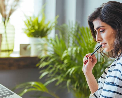 Woman Sitting at desk in striped shirt looking at a laptop and holding a pen.