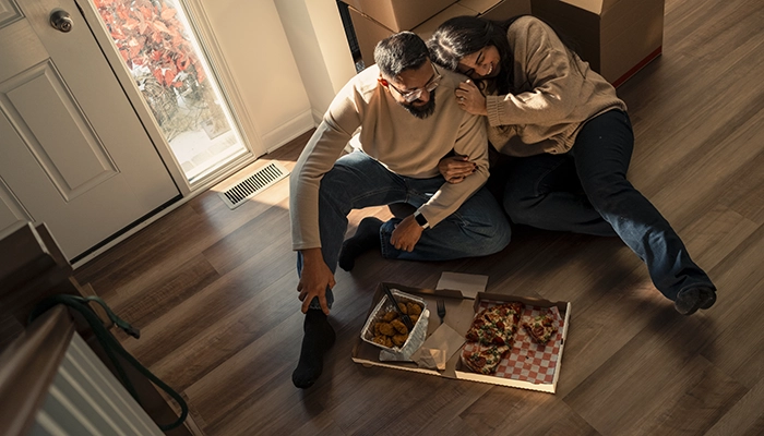 Couple laughing and sitting on floor eating pizza and chicken wings.