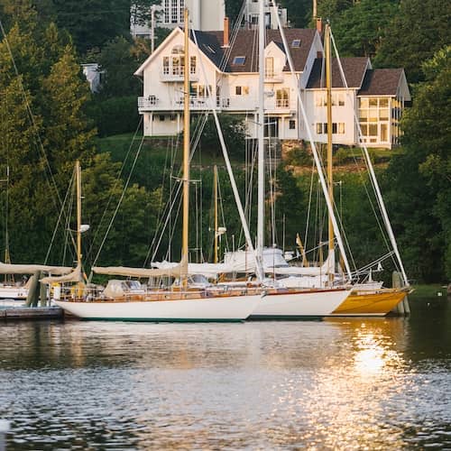 View of large white home on shore of Rockland Harbor Maine with sailboats docked nearby in the water.