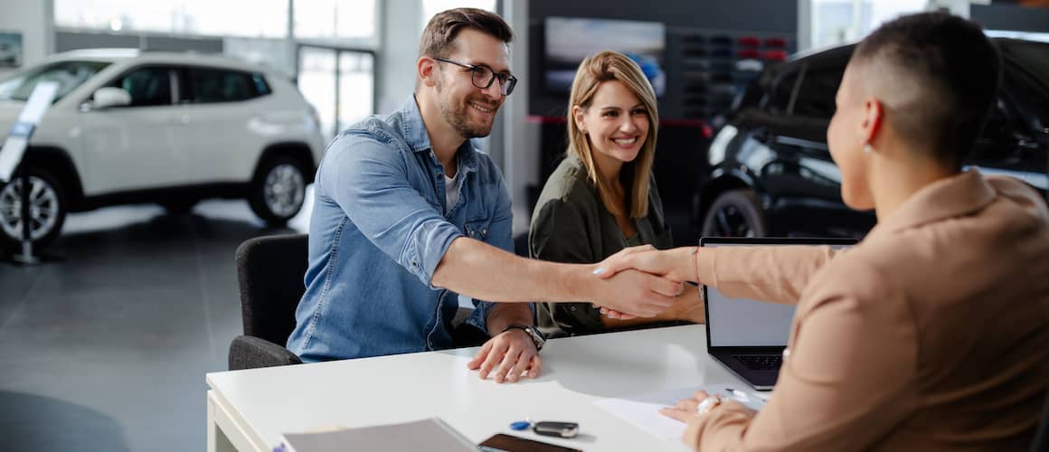 Man and woman sitting in car dealership, shaking hands with salesperson.
