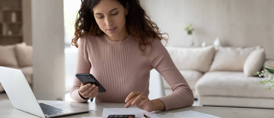 A young woman sitting at a desk, possibly managing bills or finances using a digital tablet.