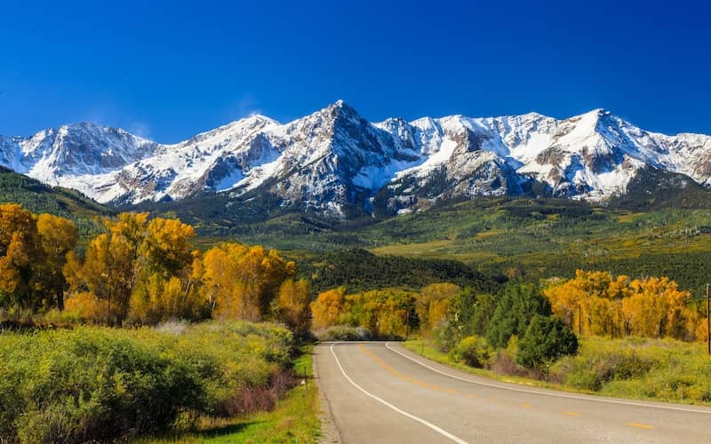 Paved road in Colorado with snow covered mountains in the background and a clear blue sky. 