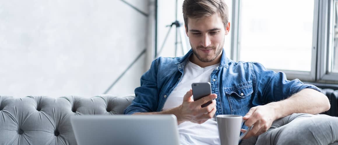 Man on couch with open laptop holding coffee cup and looking at mobile phone.