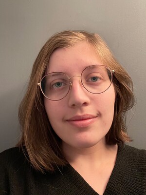 Headshot of Kit Wakelin, woman with brown blonde hair and glasses standing in front of a grey background.