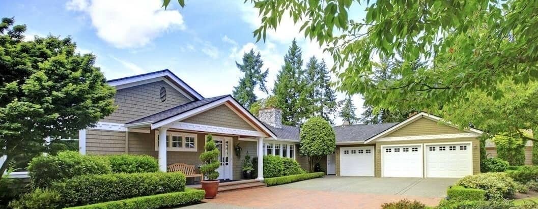 Two-story beige house with large paved driveway, manicured shrubs and red front door.