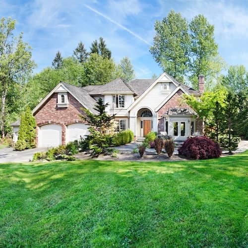 A large brick home with by woods in the background and beautiful landscaping in the front.