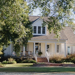 A large white house with a front porch and dormer windows sits behind a green lawn, framed by leafy trees in a quiet, sunlit neighborhood.