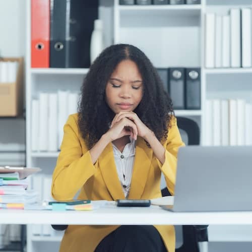 A young woman in a yellow suit coat resting her chin on her hands while deep in thought at her desk.