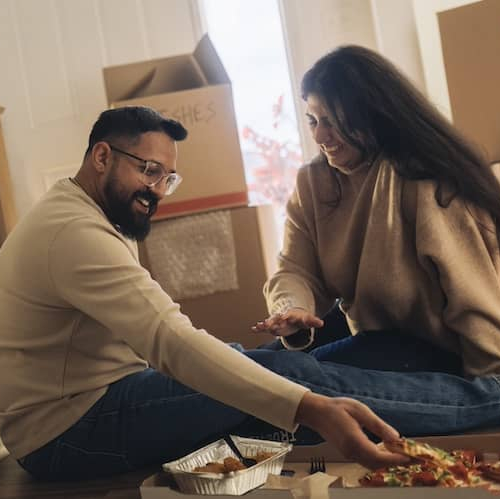 Couple smiling and eating pizza on floor of new home surrounded by unopened boxes.