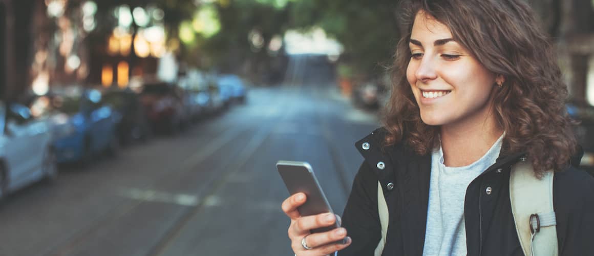 Image of woman smiling, looking down at phone with city street behind her.