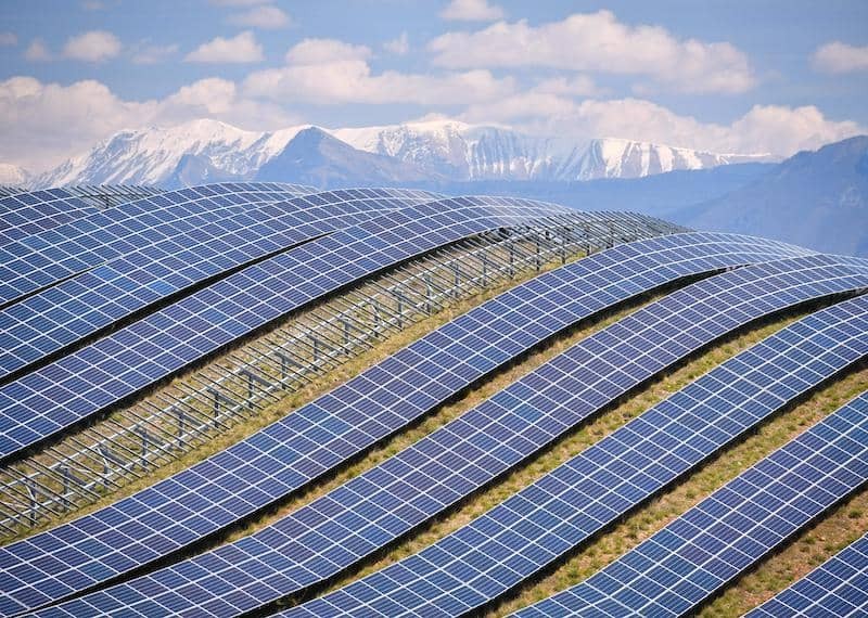 Field of photovoltaic panels on hills with snow capped mountains in the background.