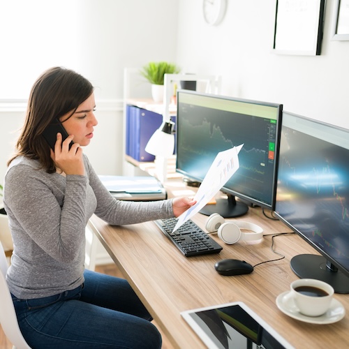 Woman looking at paperwork while on phone, sitting at desk with two computer monitors.
