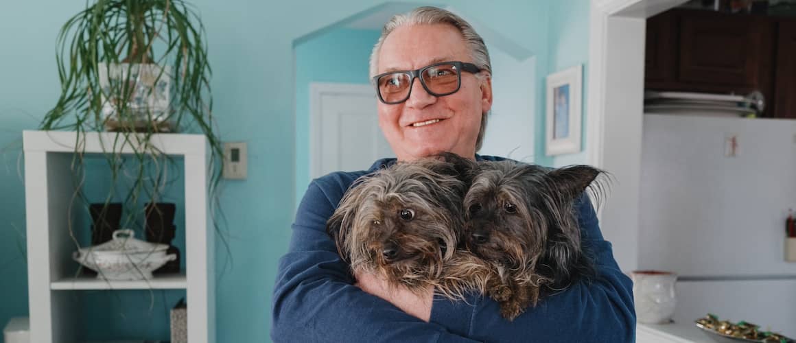 Man smiling and holding two fluffy small dogs in living room.