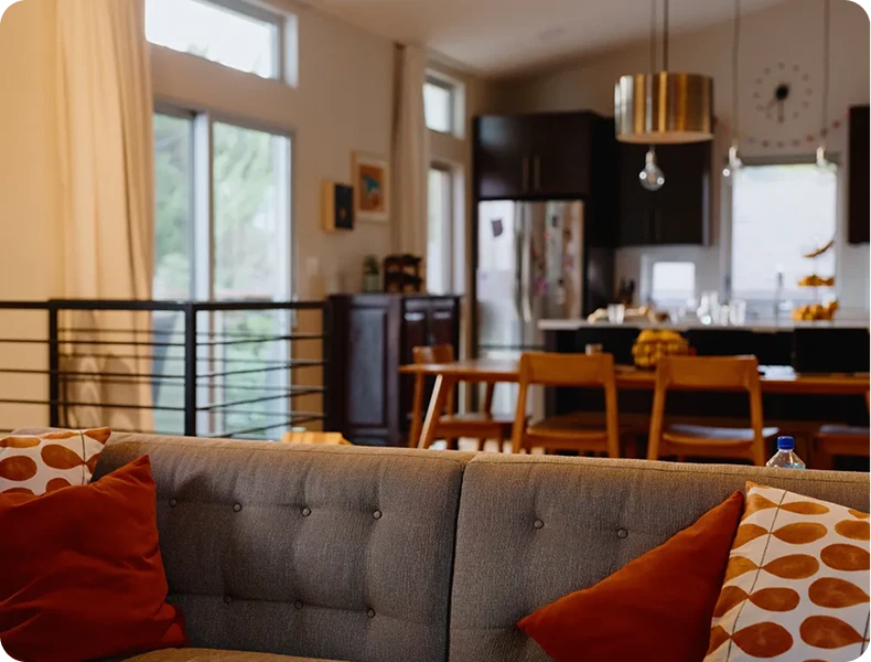 Sunlit living room with a gray sofa and colorful pillows, with a kitchen and dining area in the background.
