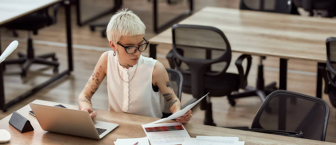 Young woman with tattoos and short white hair working on a laptop with paperwork, indicating financial or work-related tasks.