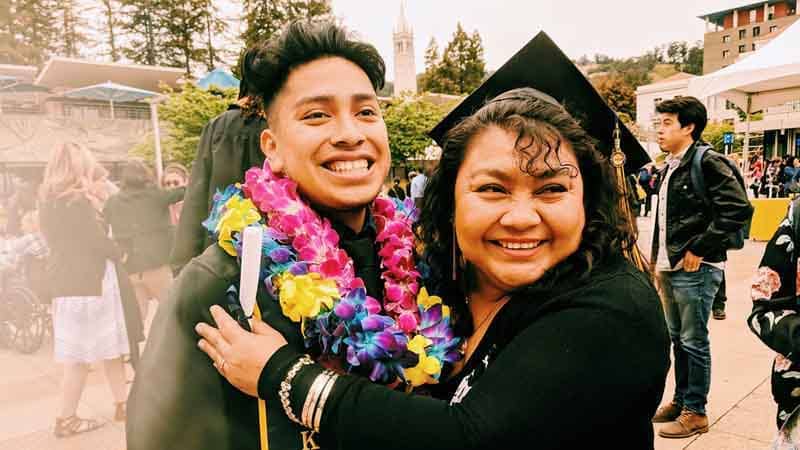 Man and his mom smiling, man in graduation robes with flower necklaces around his neck.